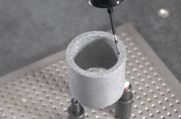 A small probe measuring the rim of a gray cylindrical part mounted above a perforated inspection table.