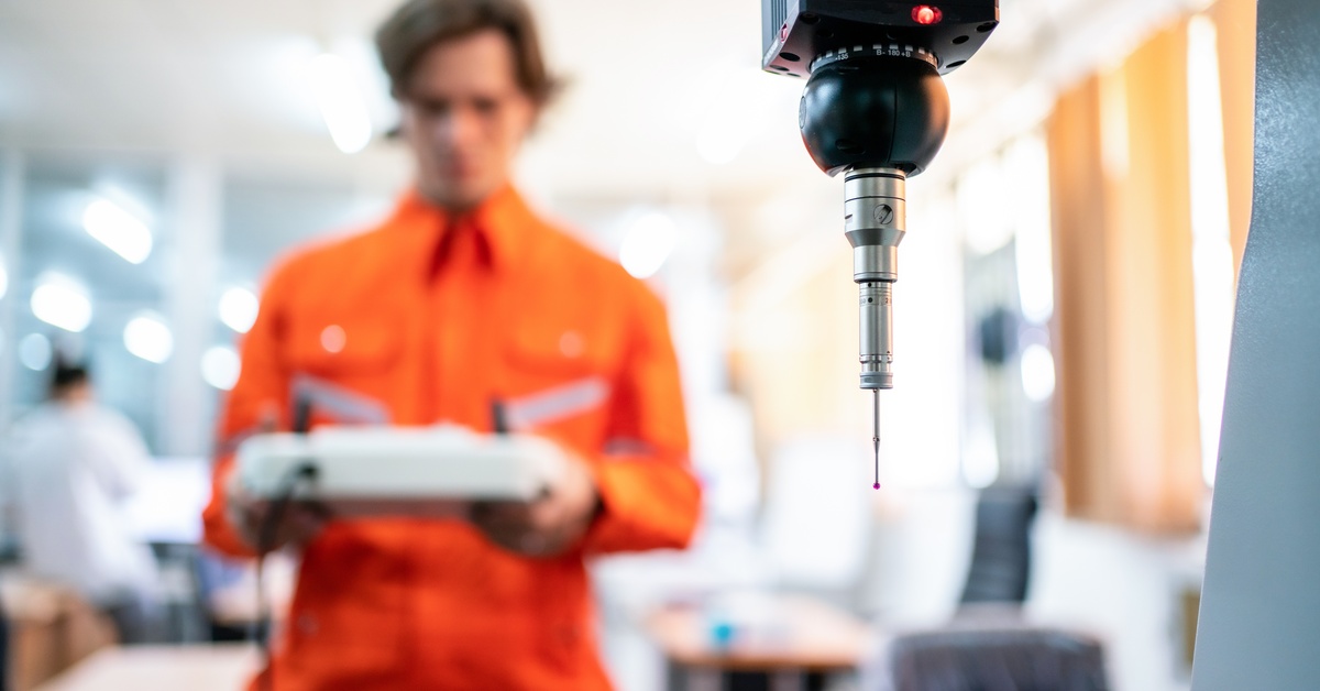 A coordinate measuring machine ready for use. A man wearing an orange jumpsuit holds the controls of the machine.