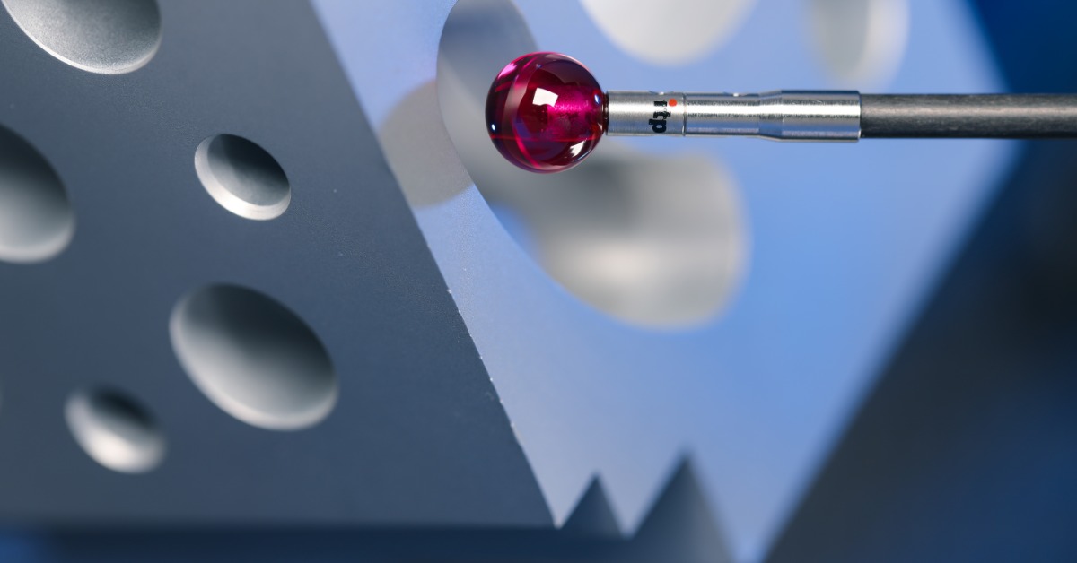A red-tipped carbon fiber stylus touching a gray block with round holes against a blue blurred background.