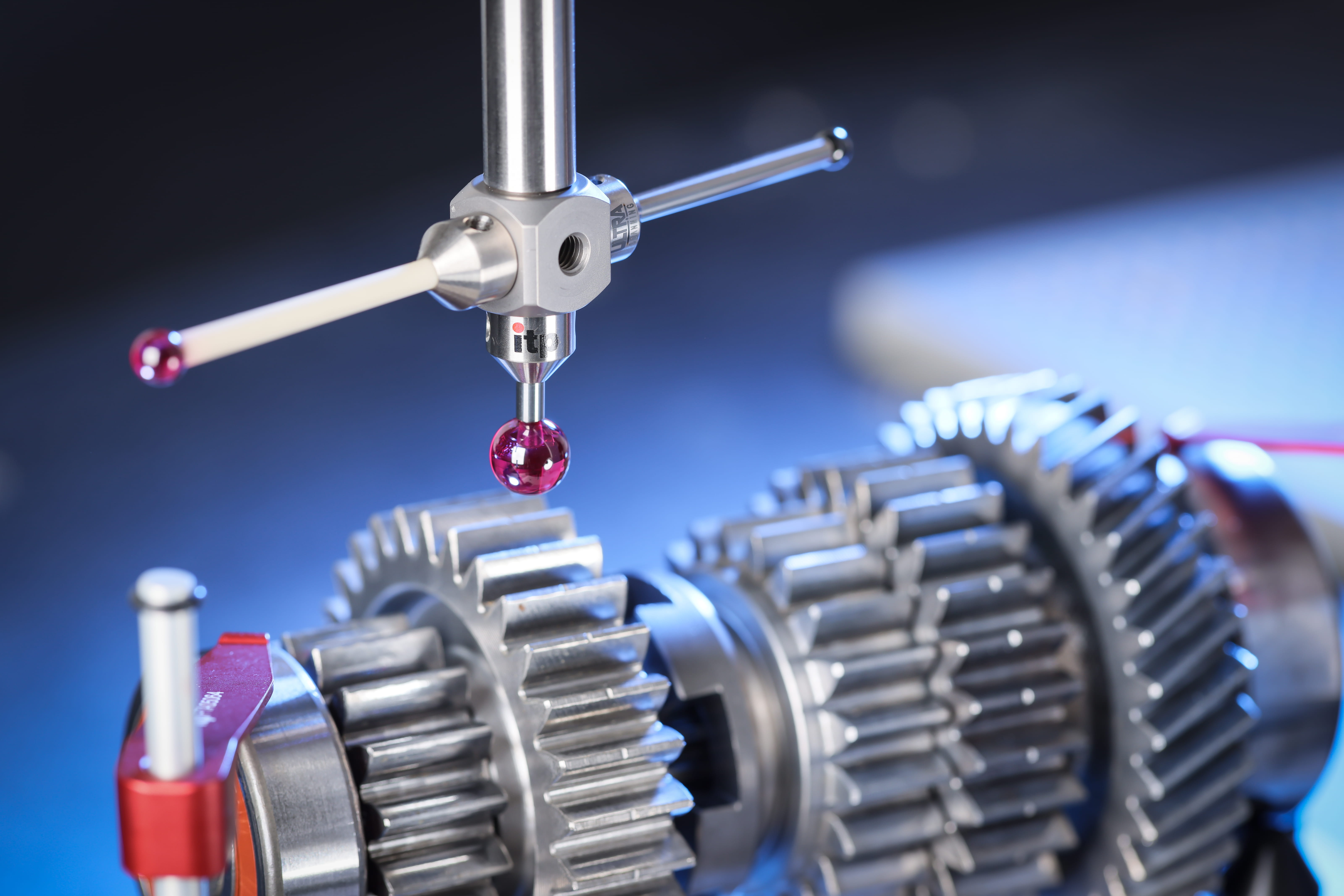 A short, ruby ball probe inspects a metal gear shaft at close range. The gear has many-sized ridges.