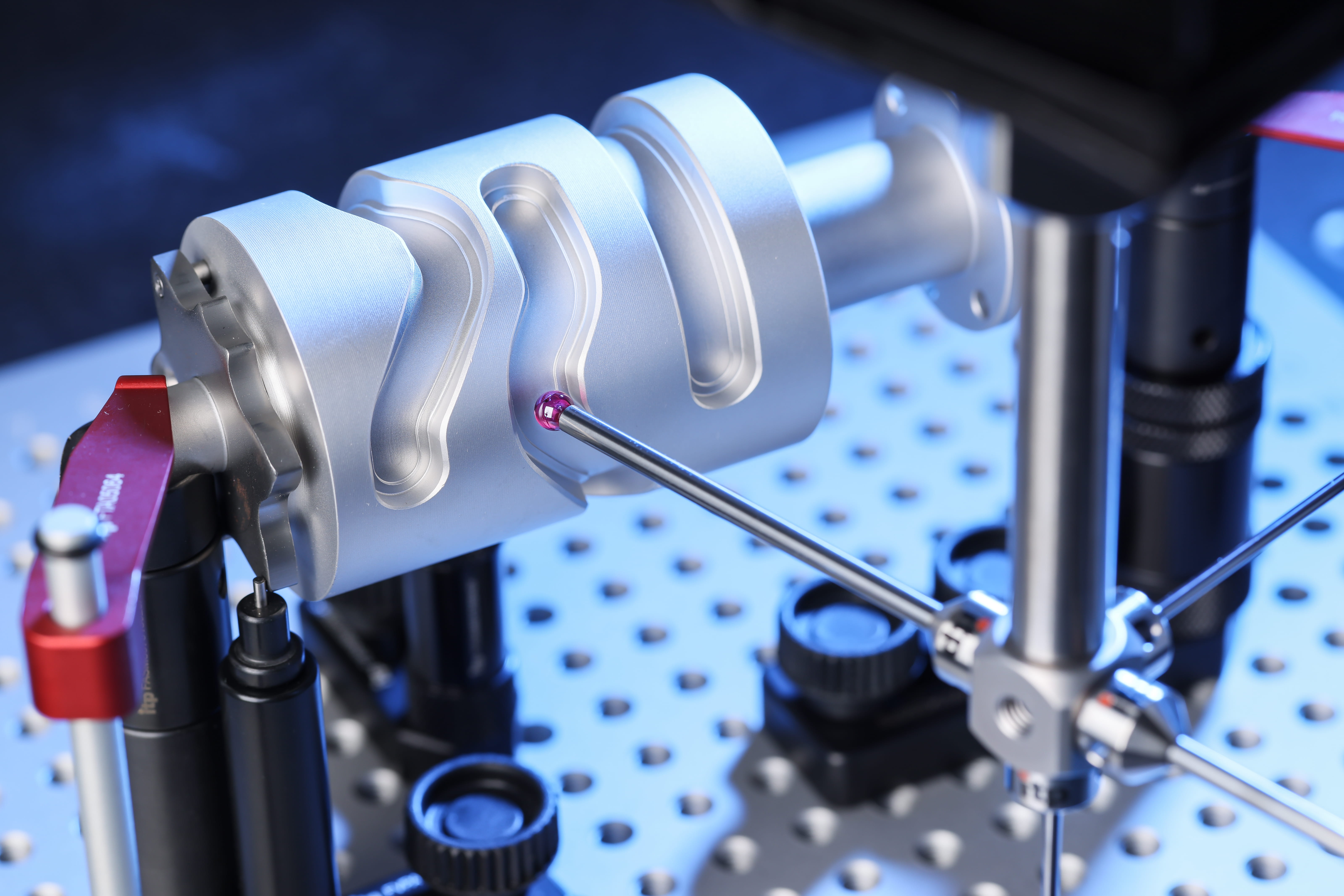 A red spherical probe touching a machined silver cylinder held on a perforated blue inspection table.