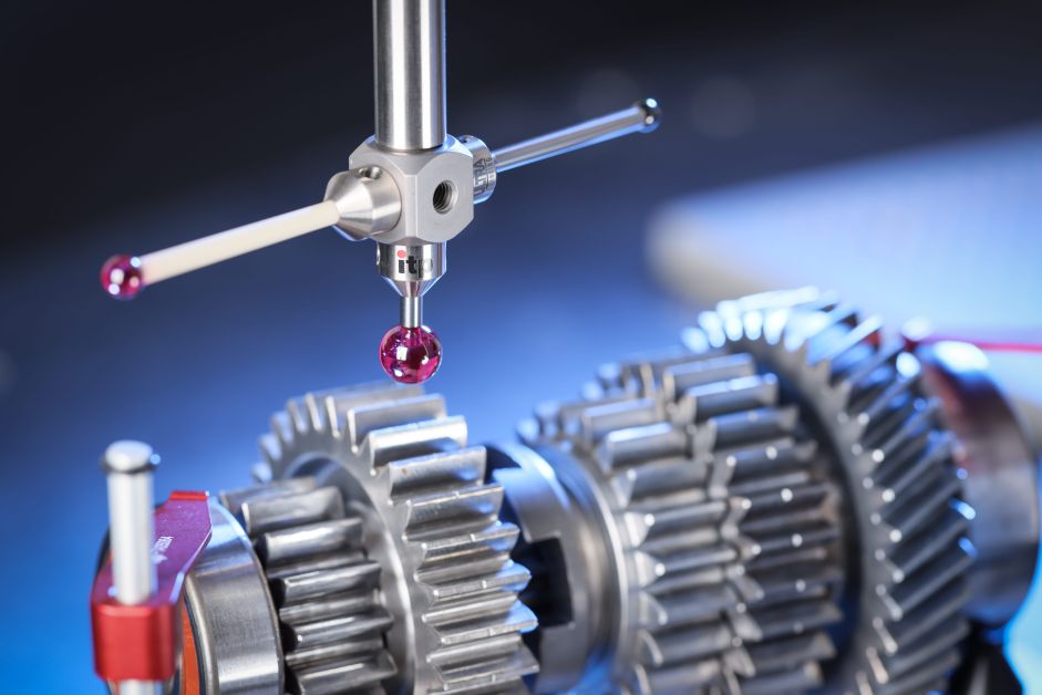 A measuring probe with red tips hovering above shiny metal gears and fixtures under blue lighting on a work surface.