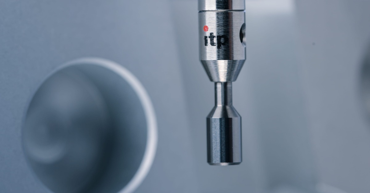 A metal measuring probe positioned near a gray machined part with circular openings against a blue background.