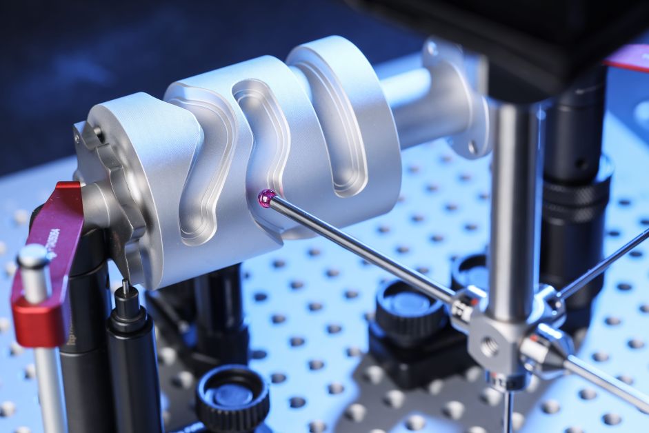 A red spherical probe touching a machined silver cylinder held on a perforated blue inspection table.
