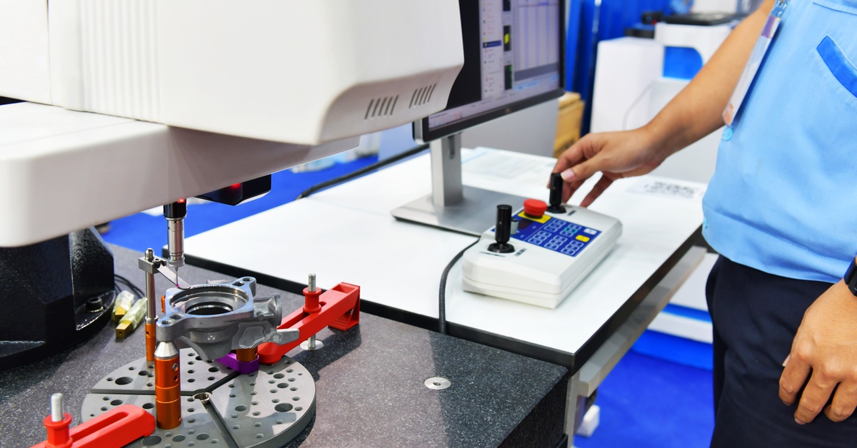 Someone stands in front of a computer with their hand on some controls. CMM equipment is working to measure a part.