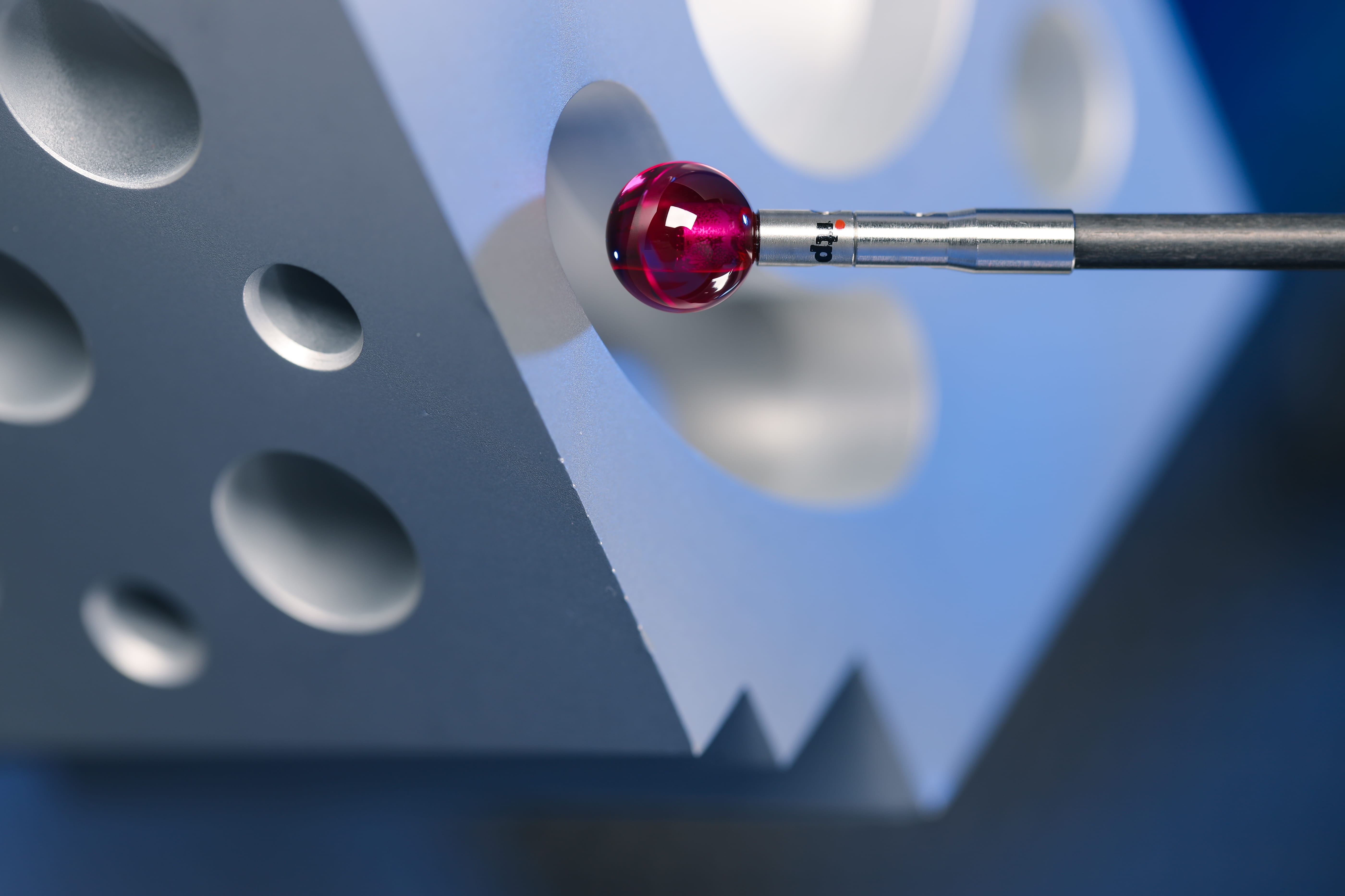 An extreme close-up of a red spherical probe touching a silver metal block with round holes and angular cutouts.