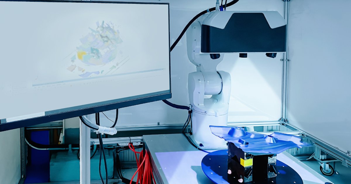 A CMM machine rests on a table next to a screen. A light shines on a part in the machine with a 3D model on the computer.