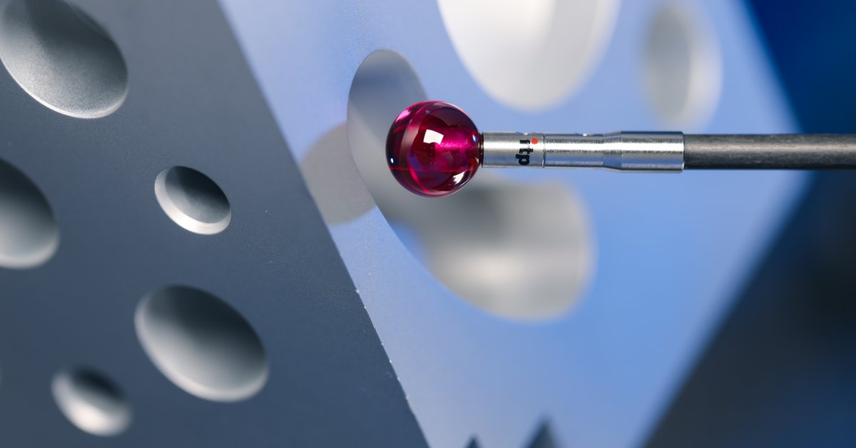 A shiny red probe tip extending from a metal rod toward circular holes in a gray machined metal surface.